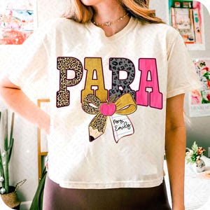May include: Cream-coloured t-shirt with the word "PARA" in a bold, colourful design. The letter "P" has a leopard print, "A" is pink, and "R" has a grey leopard print. A bow, apple, and pencil graphic is below the word.