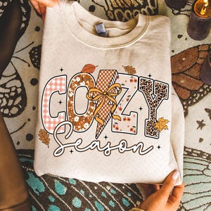 以下が含まれることがあります： 秋をテーマにしたデザインで「COZY SEASON」の文字がプリントされたクリーム色のスウェットシャツ。文字はチェック柄、花柄、ヒョウ柄、リボン付きの稲妻で装飾されています。秋の葉とさくらんぼがデザインを彩ります。