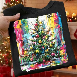 Puede incluir: Camiseta negra de cuello redondo con un diseño de árbol de Navidad abstracto y colorido. El árbol está adornado con adornos rojos, azules y amarillos. El fondo tiene un vibrante efecto de salpicaduras de pintura con rayas rosas, amarillas y azules.