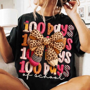 Pode incluir: T-shirt preto com a frase bordada "100 dias de escola". O design inclui um la&ccedil;o com estampa de leopardo, l&aacute;pis e padr&otilde;es de xadrez em rosa, laranja e branco. Uma pe&ccedil;a casual para o dia a dia.