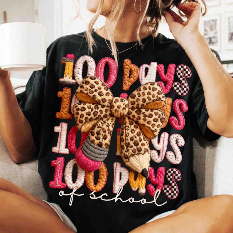 Pode incluir: T-shirt preto com a frase bordada "100 dias de escola". O design inclui um la&ccedil;o com estampa de leopardo, l&aacute;pis e padr&otilde;es de xadrez em rosa, laranja e branco. Uma pe&ccedil;a casual para o dia a dia.
