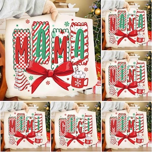 May include: A collection of Christmas-themed throw pillows with festive designs. Each pillow features a name like "MAMA," "NANA," or "MIMI" in red and green, with a large red bow, snowflakes, and a cocoa mug.