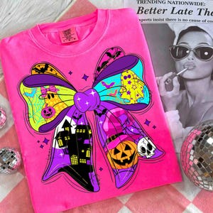 May include: A bright pink Comfort Colors sweatshirt featuring a large, colorful bow design with Halloween-themed illustrations. The bow includes images of ghosts, bats, a haunted house, a jack-o'-lantern, and a skull. The design is outlined in black and has purple, yellow, and blue accents.