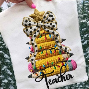 May include: A white towel with a crocheted Christmas tree design. The tree is made of yellow pencils with a black and white polka dot bow, a gold star, and colorful lights. The word "Teacher" is written in black cursive.