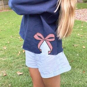 Puede incluir: Una sudadera azul marino con un lazo de cuadros rojos y blancos en la manga. La sudadera se combina con pantalones cortos de rayas azules.