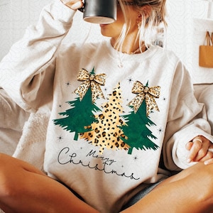 Op de afbeelding: Een wit sweatshirt met een kerstboomontwerp. De bomen zijn groen en versierd met luipaardprint strikken. De tekst "Merry Christmas" is geschreven in een cursief schrift onder de bomen.