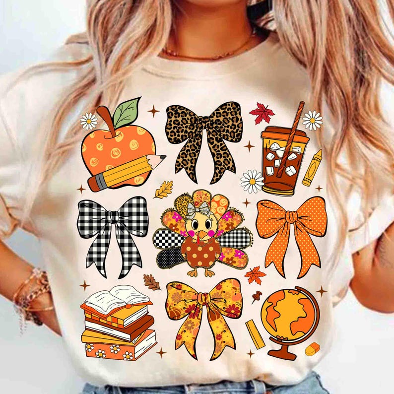 May include: A beige t-shirt featuring a collection of fall-themed illustrations. The design includes an apple with a pencil, bows, a turkey, a coffee cup, books, a globe, and fall leaves. The illustrations are in various colors, including orange, brown, and black.