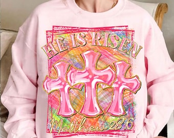 May include: Pink sweatshirt with a graphic print of three pink crosses with a rainbow background. The text "He is Risen" and "Matthew 28:6" is printed on the sweatshirt.