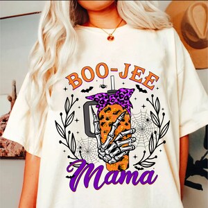 May include: White t-shirt with a graphic of a skeleton hand holding a boo-jee mama cup with a purple bandana. The cup is filled with orange and yellow liquid. The graphic is surrounded by leaves, stars, and spiderwebs.