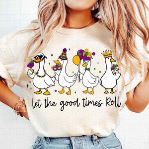 May include: Cream-colored t-shirt featuring a whimsical design of four cartoon geese dressed in Mardi Gras attire. The geese are holding accessories like masks, beads, and balloons. The phrase "let the good times Roll" is written below.