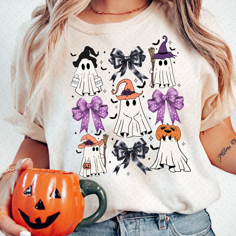 Puede incluir: Una camiseta blanca con un dise&ntilde;o de Halloween con varios fantasmas que llevan sombreros de bruja y sostienen calabazas y escobas. Los fantasmas est&aacute;n rodeados de lazos morados y negros.