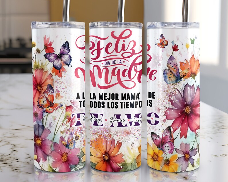 May include: A white tumbler with a floral design and butterflies. The tumbler has the text "Feliz Dia de la Madre" and "A la mejor mama' de todos los tiempos" in pink and red lettering. The tumbler also has the names "Diego", "Daniel", and "Sara" in purple lettering.