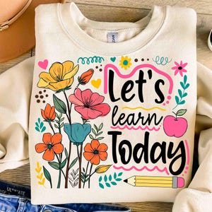 May include: Cream-colored sweatshirt with a colorful floral design and the text "Let's Learn Today." The design includes orange, yellow, pink, and blue flowers, a pink apple, and a yellow pencil.