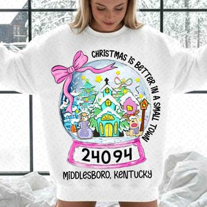 May include: White sweatshirt featuring a snow globe design with a Christmas theme. The snow globe includes a church, snowman, and reindeer. Text reads "Christmas is better in a small town" and "24094 Middlesboro, Kentucky". A pink bow is at the top.