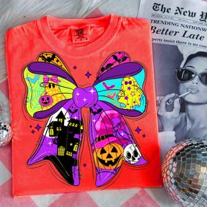 May include: Bright orange t-shirt featuring a large, colorful Halloween bow design. The bow includes ghosts, a haunted house, a witch's hat, and a jack-o'-lantern. The shirt has a tag that reads "Comfort Colors". A newspaper with the headline "Better Late" is in the background.