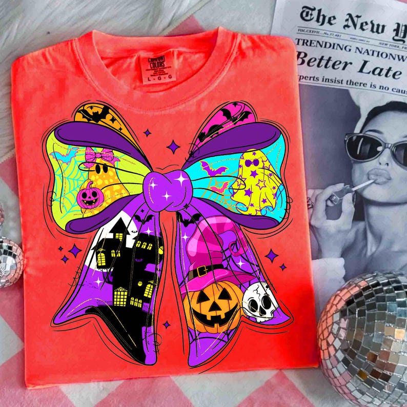 May include: Bright orange t-shirt featuring a large, colorful Halloween bow design. The bow includes ghosts, a haunted house, a witch's hat, and a jack-o'-lantern. The shirt has a tag that reads "Comfort Colors". A newspaper with the headline "Better Late" is in the background.