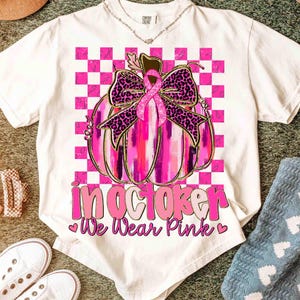 May include: White t-shirt featuring a pink pumpkin with a leopard print bow and a pink ribbon. The shirt has the text "In October We Wear Pink" in pink lettering. The design is set against a pink and white checkered background.
