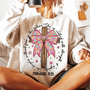 May include: A white sweatshirt featuring a pink and brown cross with a bow. The text on the sweatshirt reads "She laughs without fear of the future + She is clothed with strength + dignity PROVERBS 31:25".