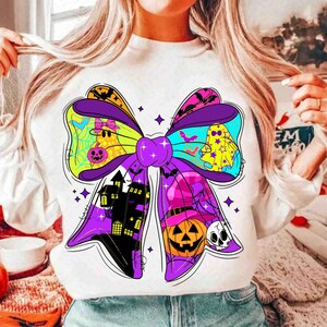 May include: White sweatshirt featuring a large, colorful Halloween bow design. The bow includes a haunted house, jack-o'-lantern, ghosts, bats, and spiderwebs. The bow is outlined in black and has purple, green, yellow, and blue accents.