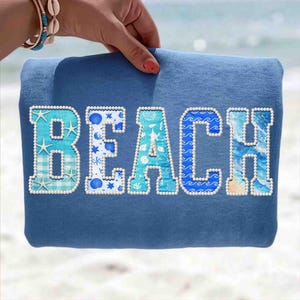 May include: A blue sweatshirt with the word "BEACH" appliqued across the front. The letters are decorated with various beach-themed patterns, including starfish, waves, and seashells, and are outlined with small white beads.