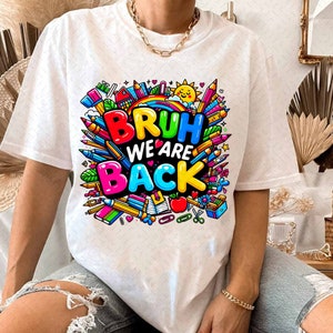 Puede incluir: Camiseta blanca con un dise&ntilde;o gr&aacute;fico colorido que presenta el texto "BRUH WE ARE BACK" rodeado de &uacute;tiles escolares como l&aacute;pices, crayones y una manzana.