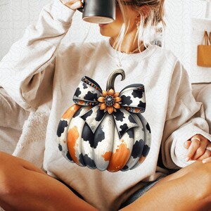 May include: A white sweatshirt with a black and orange pumpkin with a cow print bow. The pumpkin has a brown stem and a black and white flower with a rhinestone center.