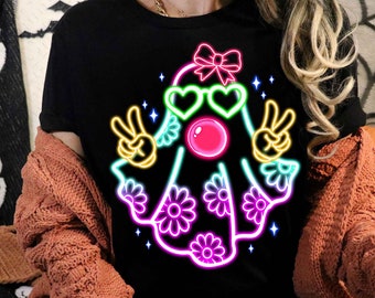 May include: A black t-shirt featuring a neon pink, green, and yellow graphic of a ghost wearing heart-shaped sunglasses and a bow. The ghost is surrounded by flowers and stars.