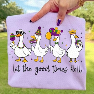 May include: A light purple t-shirt featuring four cartoon geese dressed for a Mardi Gras celebration. Each goose wears festive accessories like sunglasses, a mask, a party hat, and a crown. The text "Let the good times Roll" is written in black script.