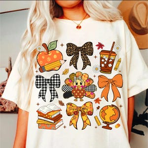 May include: Cream-colored t-shirt featuring colorful fall-themed graphics. Designs include an apple with a pencil, bows, a turkey, a coffee cup, books, a globe, and fall leaves. The shirt has a relaxed fit.