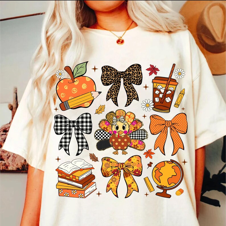 May include: Cream-colored t-shirt featuring colorful fall-themed graphics. Designs include an apple with a pencil, bows, a turkey, a coffee cup, books, a globe, and fall leaves. The shirt has a relaxed fit.