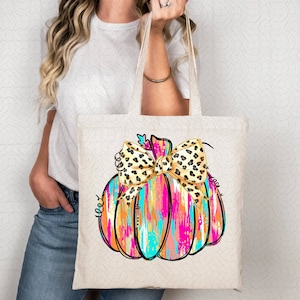 May include: A white tote bag with a colorful painted pumpkin with a leopard print bow. The pumpkin is pink, orange, blue, and gold.
