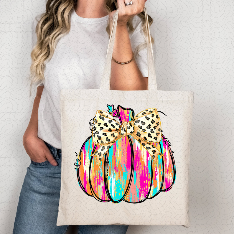 May include: A white tote bag with a colorful painted pumpkin with a leopard print bow. The pumpkin is pink, orange, blue, and gold.