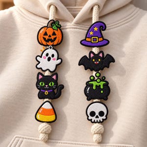 May include: A beige hoodie with two thick cream-coloured drawstrings decorated with embroidered Halloween patches. The left string features a pumpkin, ghost, black cat, and candy corn. The right string features a witch hat, bat, cauldron, and skull.