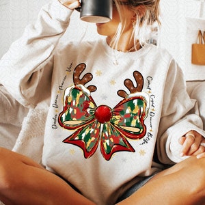May include: White sweatshirt with a red and green camouflage bow with gold glitter accents. The bow has a red pom-pom in the center and reindeer antlers on either side. The text "Dasher Dancer Prancer Vixen Comet Cupid Donner Blitzen" is printed on the sweatshirt.