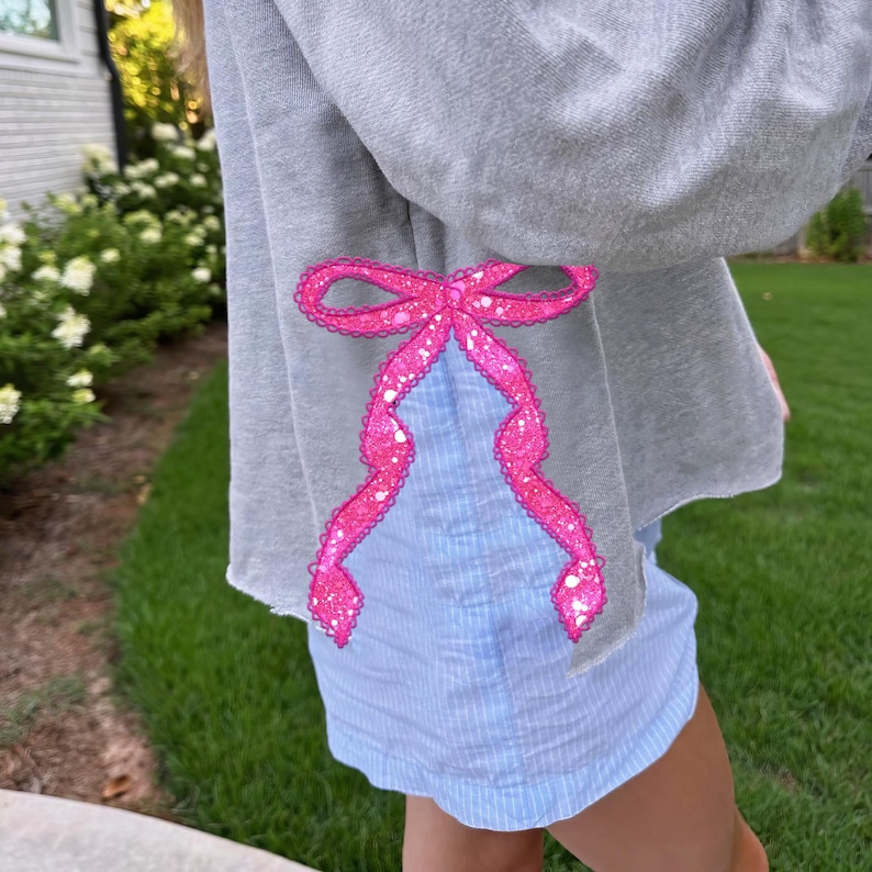 Puede incluir: Una sudadera gris con un lazo rosa brillante cosido en la manga. La sudadera se lleva sobre una camisa azul claro a rayas.