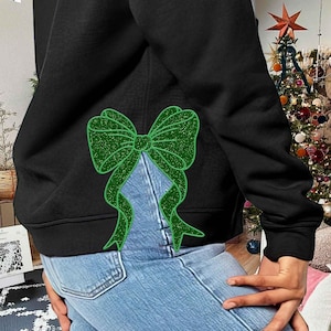 Może przedstawiać: Czarny sweterek z zieloną błyszczącą kokardką z tyłu. Sweterek jest noszony na niebieskich dżinsach.