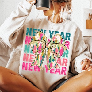 May include: A white sweatshirt with the words "NEW YEAR" repeated four times in a vintage style font. A large, colorful bow with gold glitter accents is centered on the sweatshirt.