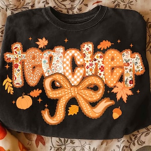 May include: Black sweatshirt with the word "teacher" in orange, decorated with fall-themed patterns, including leopard print, gingham, and floral designs. A large orange bow and autumn leaves complete the design. A pumpkin and acorn are also included.