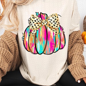 May include: A white t-shirt with a colorful abstract pumpkin design. The pumpkin has a leopard print bow on top.