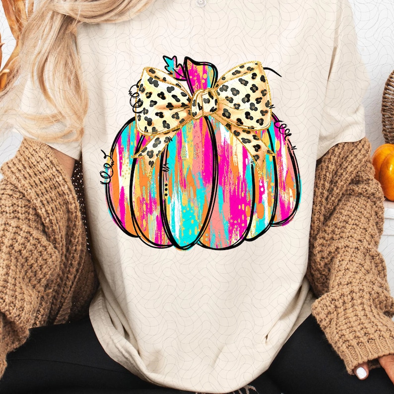 May include: A white t-shirt with a colorful abstract pumpkin design. The pumpkin has a leopard print bow on top.