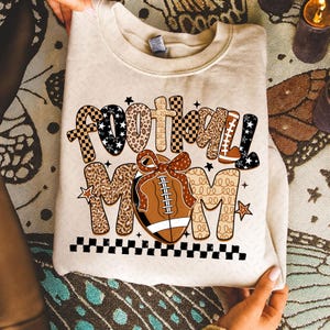 May include: Cream-colored sweatshirt with the words "football MOM" in a mix of patterns and colors. The design includes a football with a bow, stars, and a checkered border. The sweatshirt is held by hands.