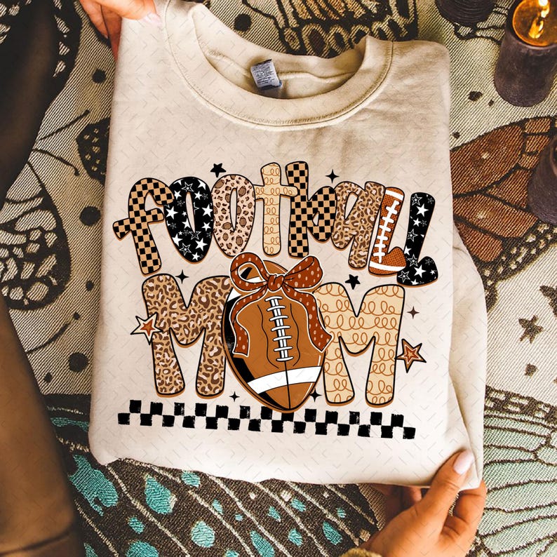May include: Cream-colored sweatshirt with the words "football MOM" in a mix of patterns and colors. The design includes a football with a bow, stars, and a checkered border. The sweatshirt is held by hands.
