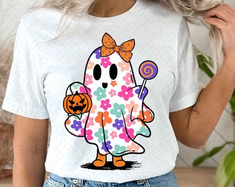 May include: A white t-shirt featuring a cute cartoon ghost wearing a pink bow and holding a pumpkin and a lollipop. The ghost is covered in colourful flowers.