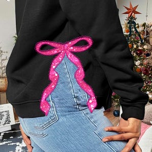 Puede incluir: Sudadera negra con un dise&ntilde;o de lazo rosa brillante en la espalda. La sudadera se lleva sobre un vaquero azul.