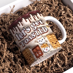 Puede incluir: Una taza de cerámica blanca con un diseño marrón, blanco y amarillo. La taza tiene un asa y dice "Let's get toasted" con un gráfico de un malvavisco, una barra de chocolate y galletas graham.