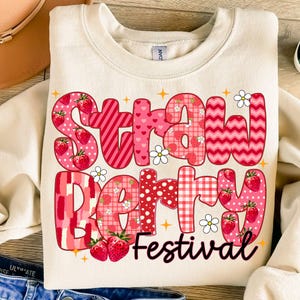 May include: A cream-colored sweatshirt with a pink and red patchwork design that says "Strawberry Festival".