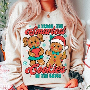 May include: A beige sweatshirt with a Christmas-themed design featuring two gingerbread people. The text on the sweatshirt reads "I teach the smartest cookies in the batch."