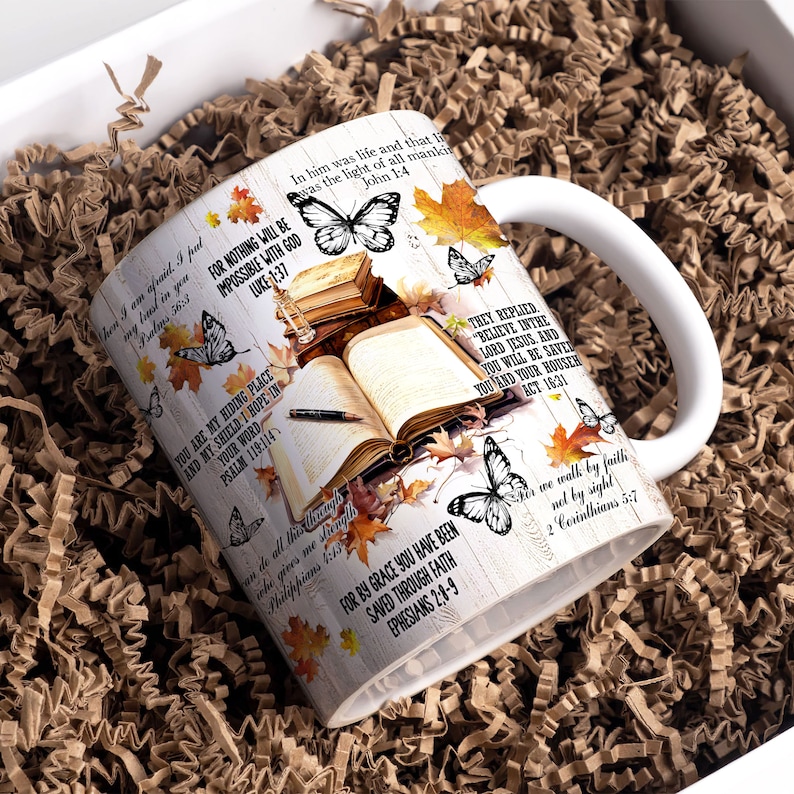May include: White ceramic mug with a colorful autumn leaf design and inspirational bible verses. The mug features butterflies, a book, and a pen. The text includes verses from Luke, Psalms, John, Philippians, Ephesians, Acts, and 2 Corinthians.