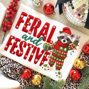May include: A white pillow with the words "FERAL and FESTIVE" embroidered in red and green. A crocheted raccoon wearing a Santa hat, scarf, and Christmas sweater holds a gift and makes a peace sign. Christmas ornaments and decor surround the pillow.