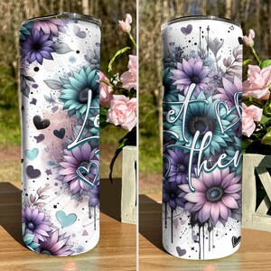 May include: A white stainless steel tumbler with a floral design featuring purple, blue, and pink flowers. The tumbler has a white background with black splatters of paint. The text "Let the" is visible on the tumbler.